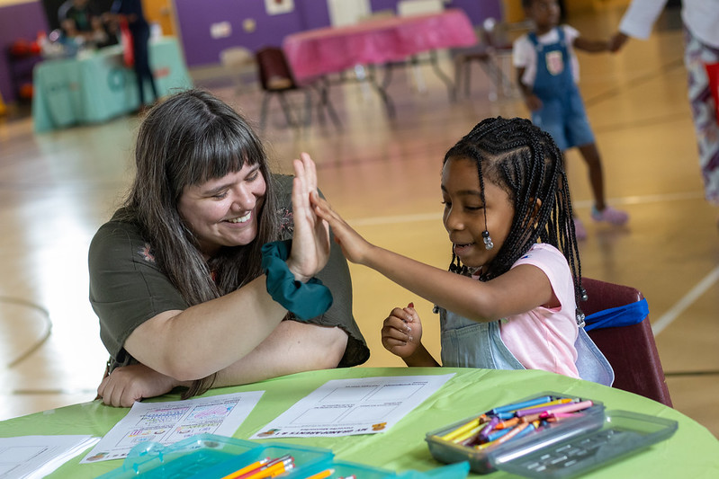 Connectedly’s Outreach Engagement Specialist gives a high five to a young girl seated at a table with drawing supplies.