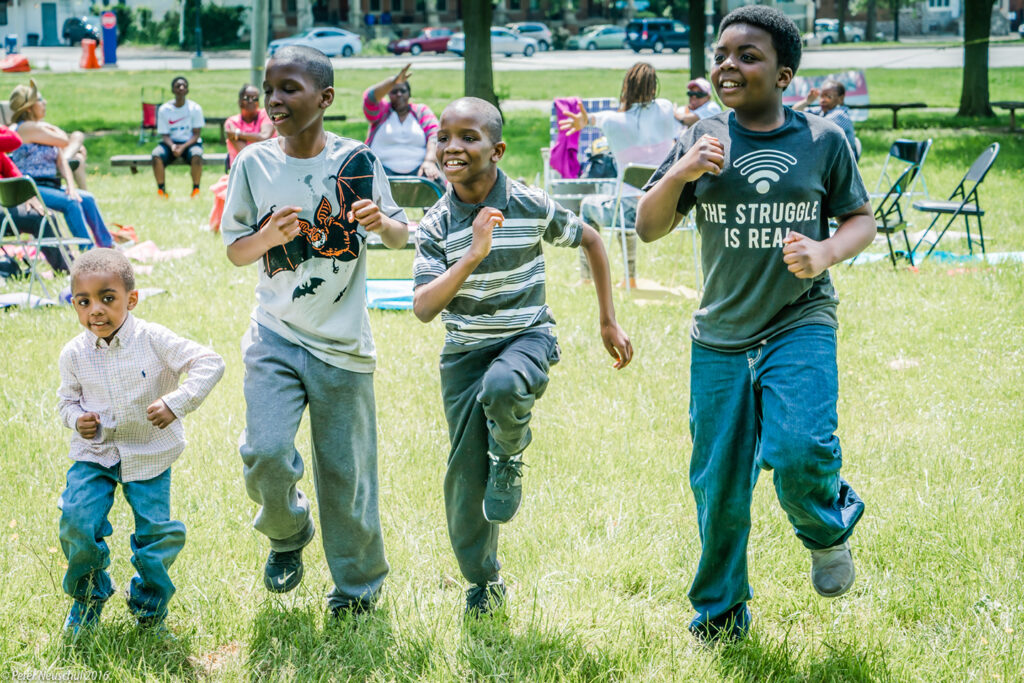 Four boys exercise at the park while their grandparent caregivers do chair yoga in the background.